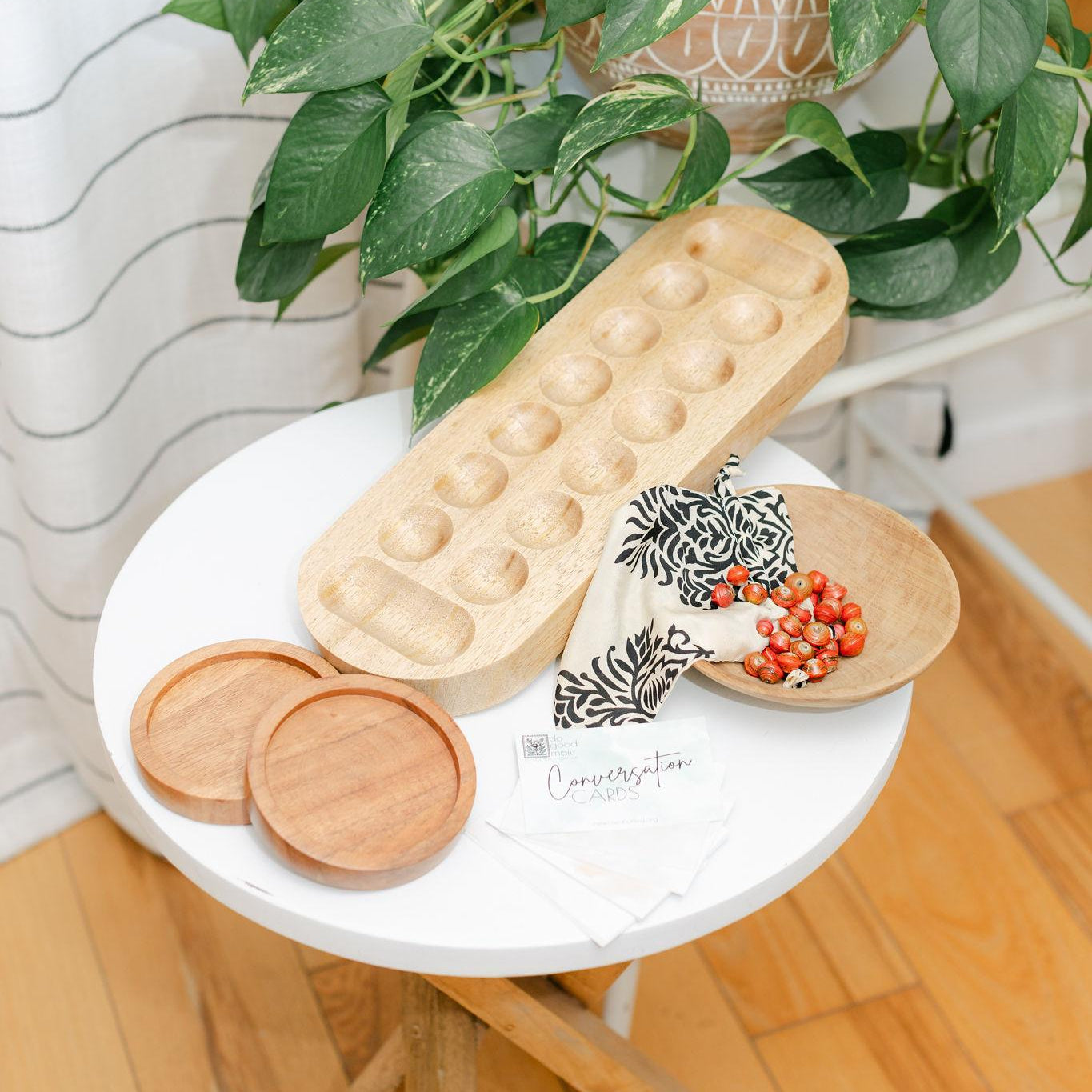 Round white table with a wooden mancala board, wooden coasters, and a plant in the background.