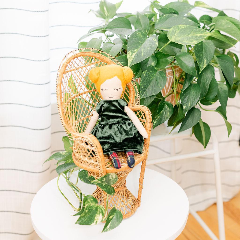 A doll with blonde hair and buns wearing a green dress, sitting in a wicker chair with a plant in the background.