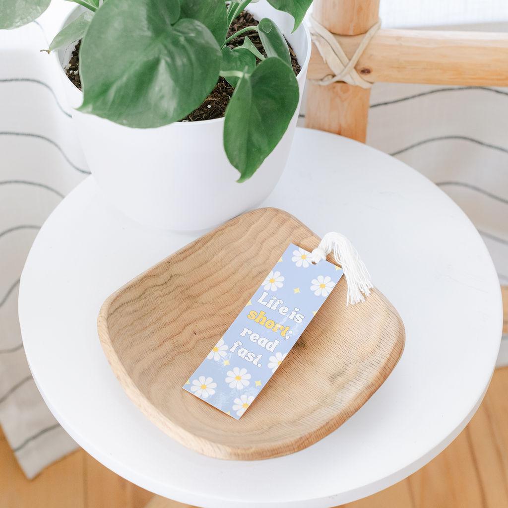 A handmade bookmark with a blue and white floral pattern resting on a wooden surface, with a white plant pot in the background.