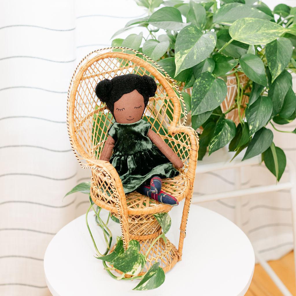 A doll with black hair and buns wearing a green dress, sitting in a wicker chair with a plant in the background.