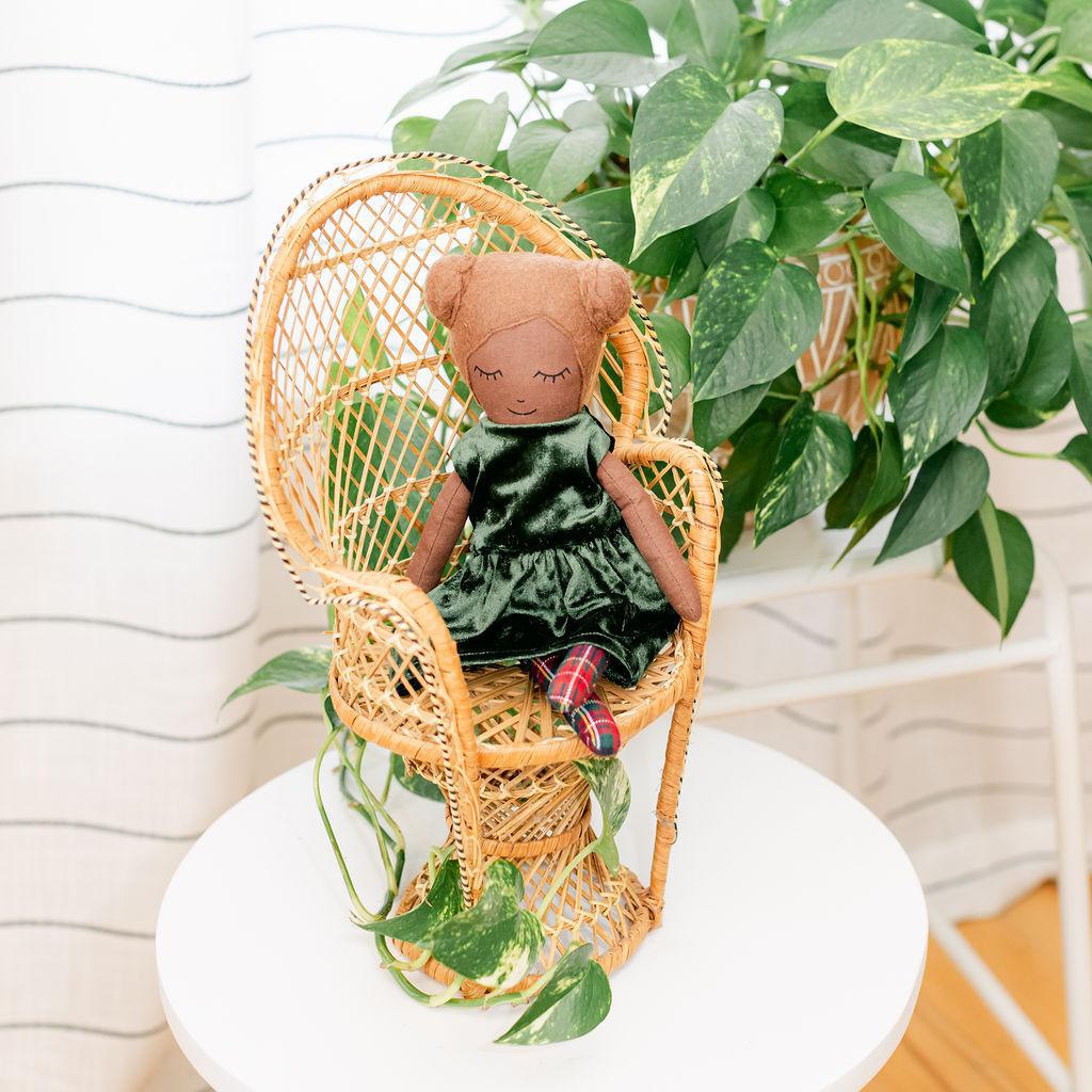 A doll with brown hair and buns wearing a green dress, sitting in a wicker chair with a plant in the background.