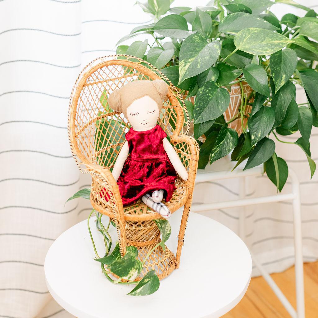 A doll with a maroon velvet dress presented sitting in a wicker chair against a backdrop of green plants.