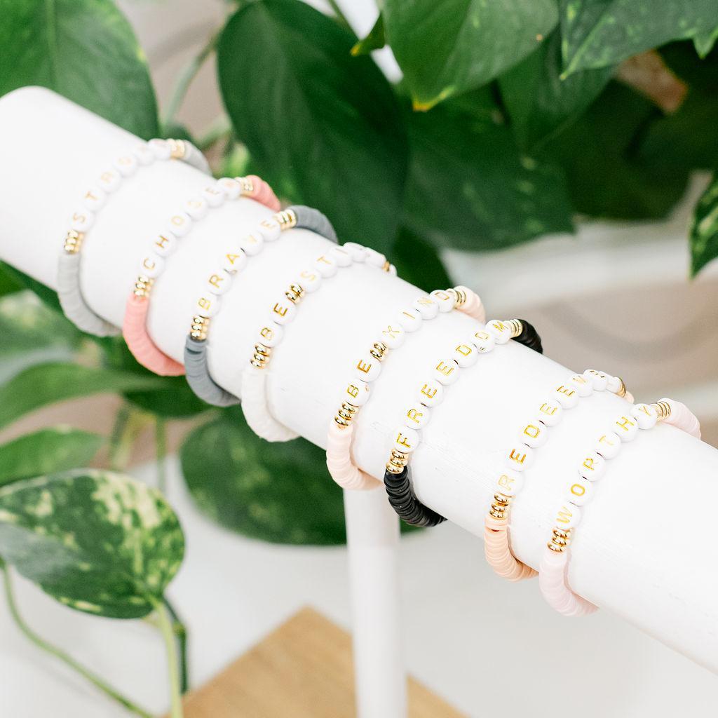 A series of bead affirmation bracelets in various colors displayed on a white stand, with each bracelet featuring different words written in a circular pattern.