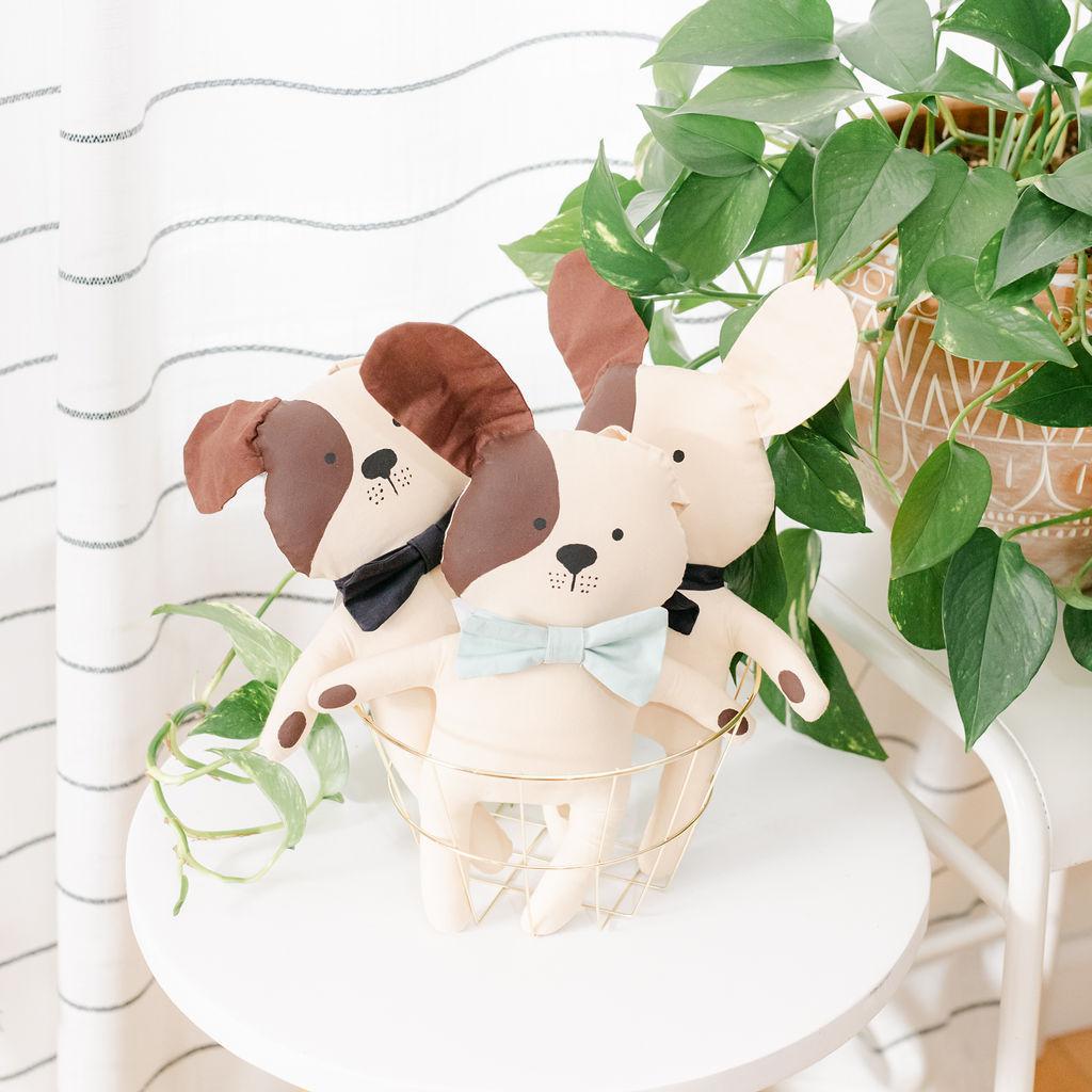 A handcrafted plush toy resembling a dog with a beige body, wearing a brown collar and a blue bow tie. It is displayed in a room with a green potted plant in the background.