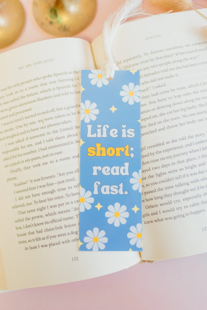 A handmade bookmark with a blue and white floral pattern resting on an open book with brass items in the background
