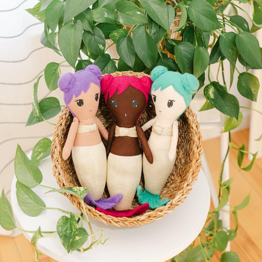 Three mermaid dolls with gold clothing in a basket with a plant in the background