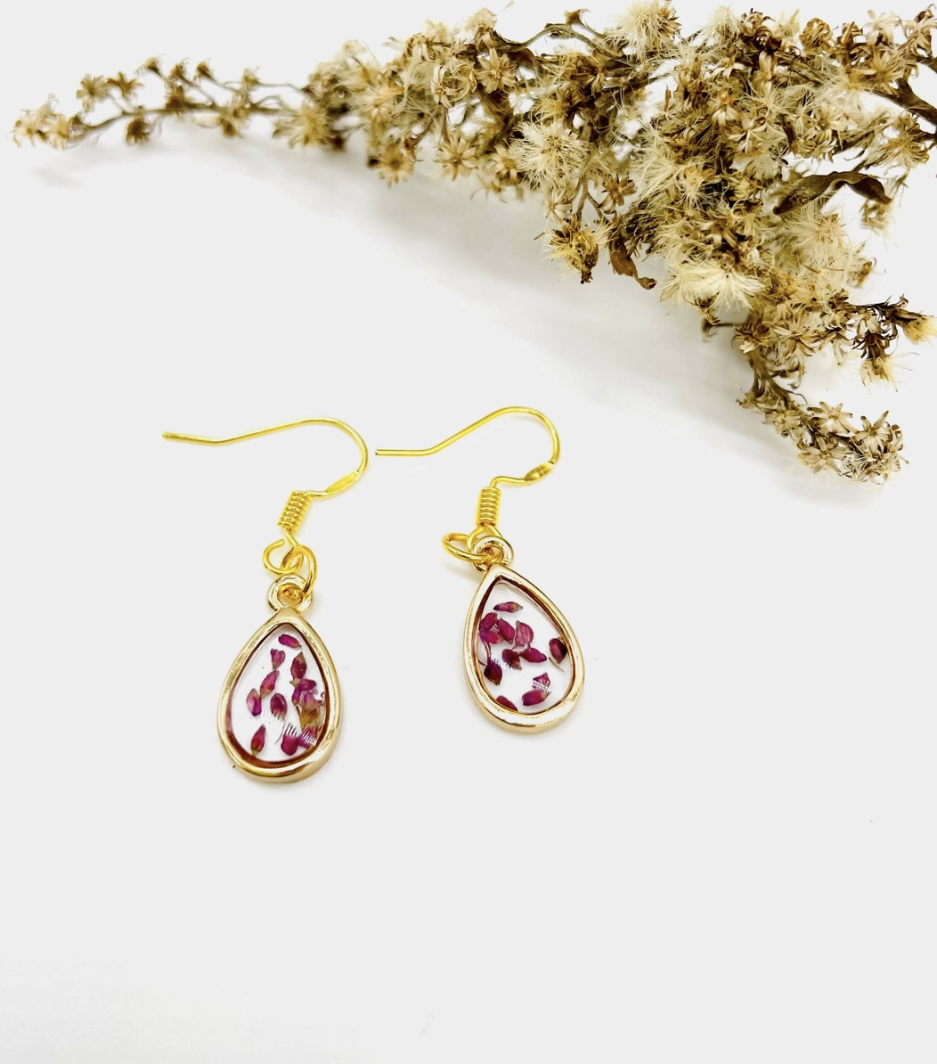 A pir of gold resin earrings with red flower pedals with flowers in the background