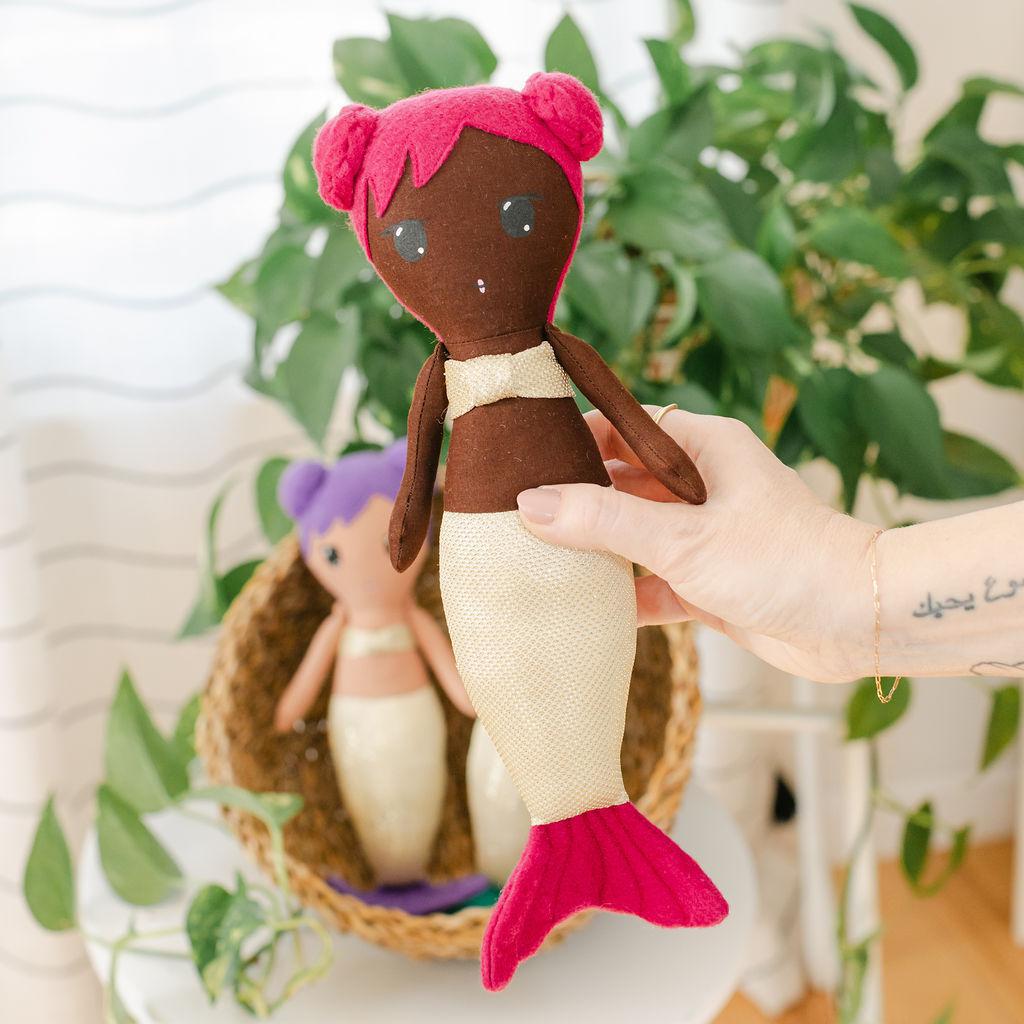 Handcrafted doll resembling a mermaid with a handpainted face, brown skin, pink buns, and a sparkly tail, held in front of a basket with a plant.