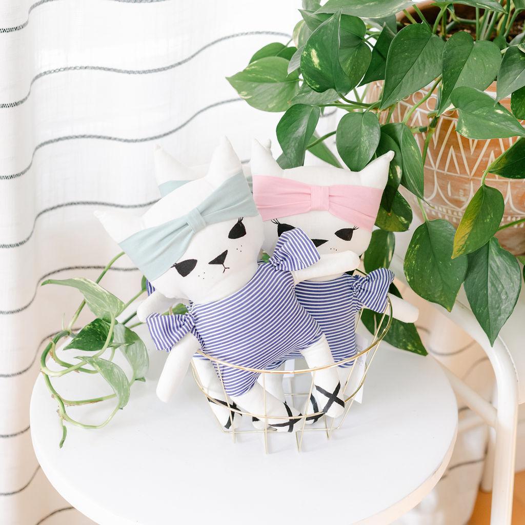 Two plush toy cats in a basket, one with a blue headband and the other with a pink headband, dressed in striped shirts.