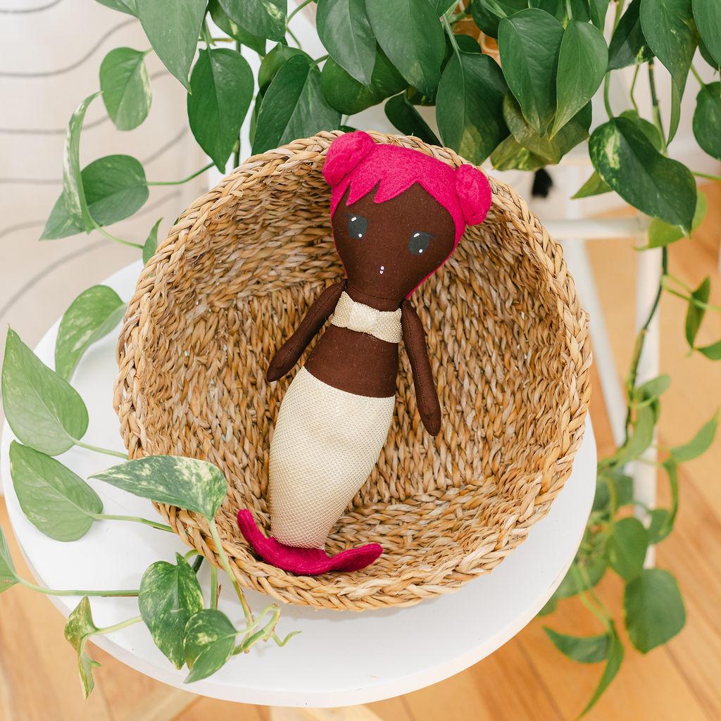 A mermaid doll with dark skin and pink hair laying in a basket on a white able with a plant in the background