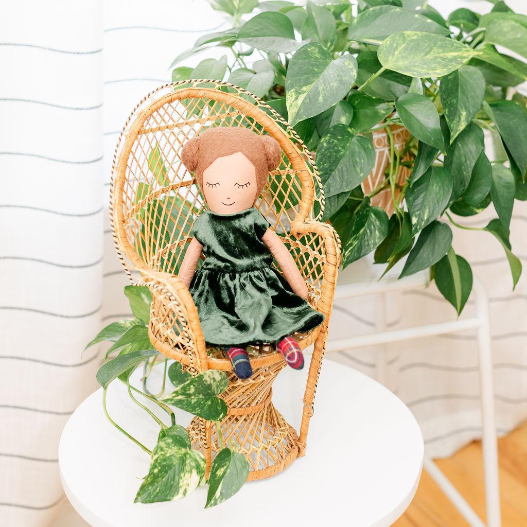 A doll with brown hair and buns wearing a green dress, sitting in a wicker chair with a plant in the background.