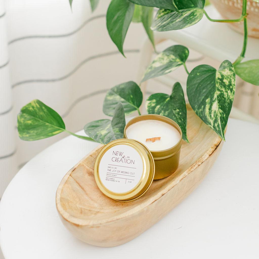 A hand-poured soy candle in a white container with a gold lid placed on a wooden surface with green leafy background.