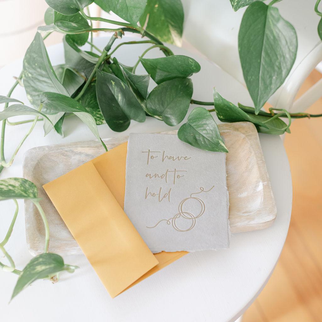 A gray wedding invitation card with gold lettering, accompanied by a light brown envelope, displayed on a white surface with green leafy branches in the background.