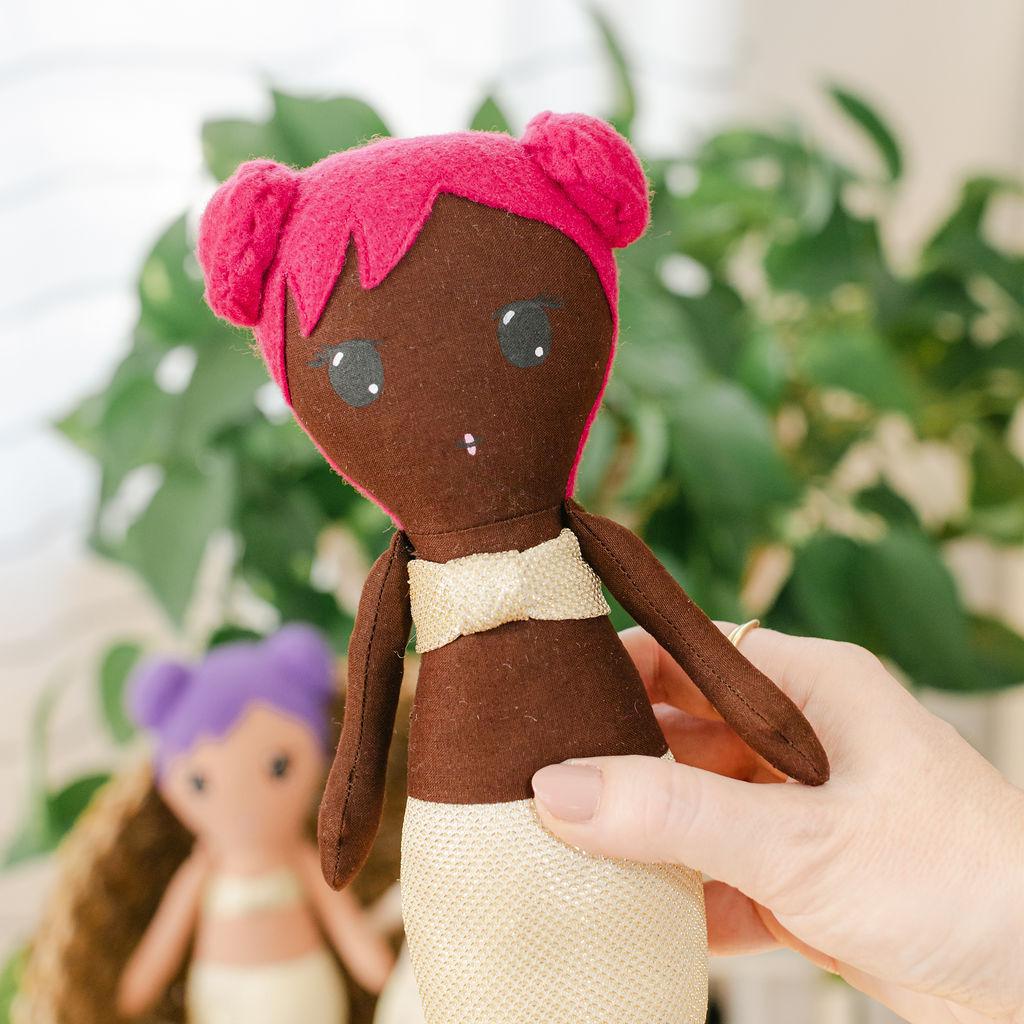 A stuffed mermaid doll with dark skin and pink hair wearing gold clothing and a plant in the background