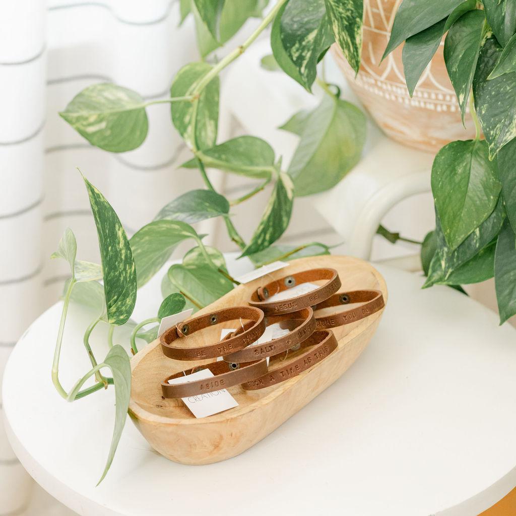Wooden dish with a collection of brown leather bracelets featuring different sayings on a white table with a plant in the background