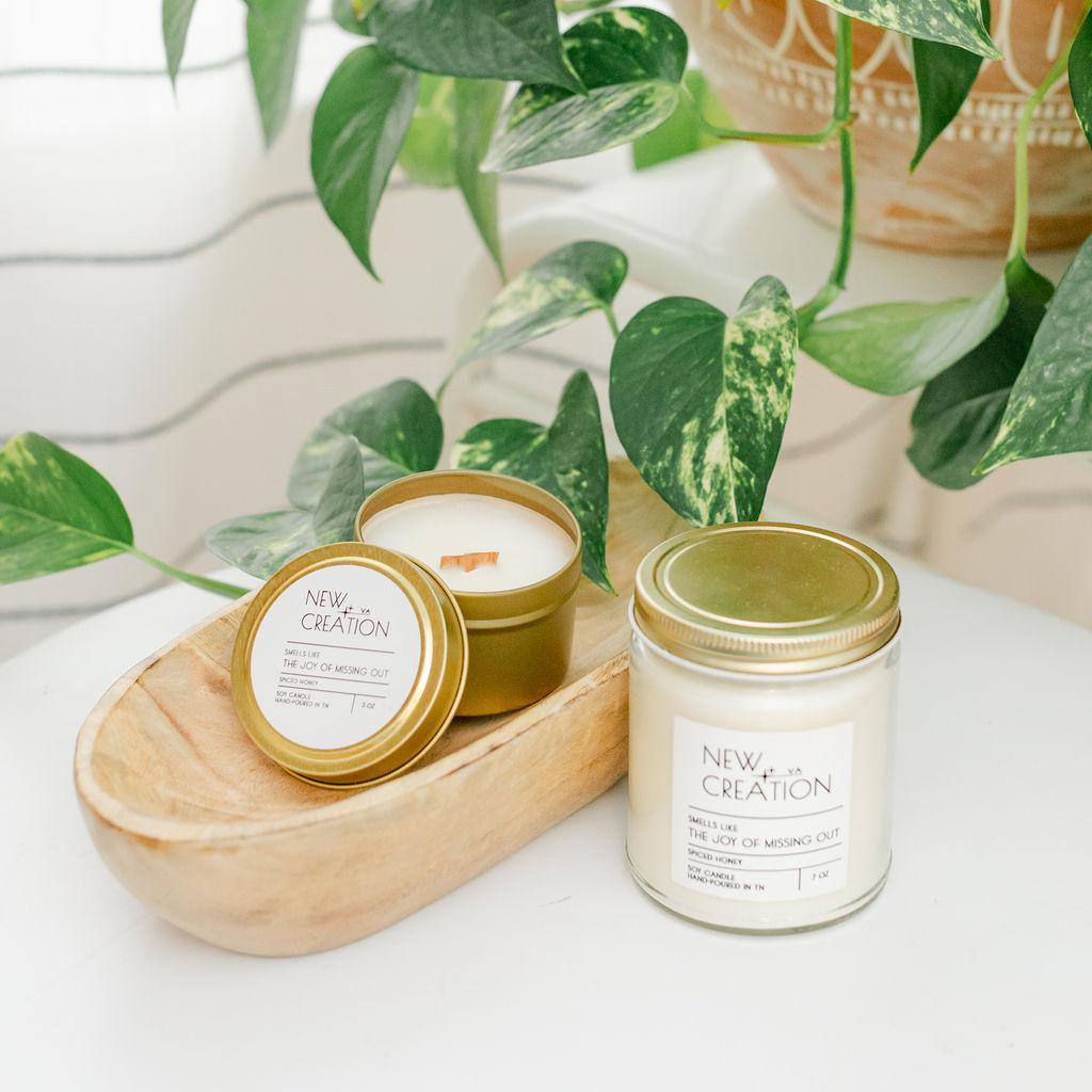 A hand-poured soy candle in a white container with a gold lid, alongside a smaller tin candle, placed on a wooden surface with green leafy background.