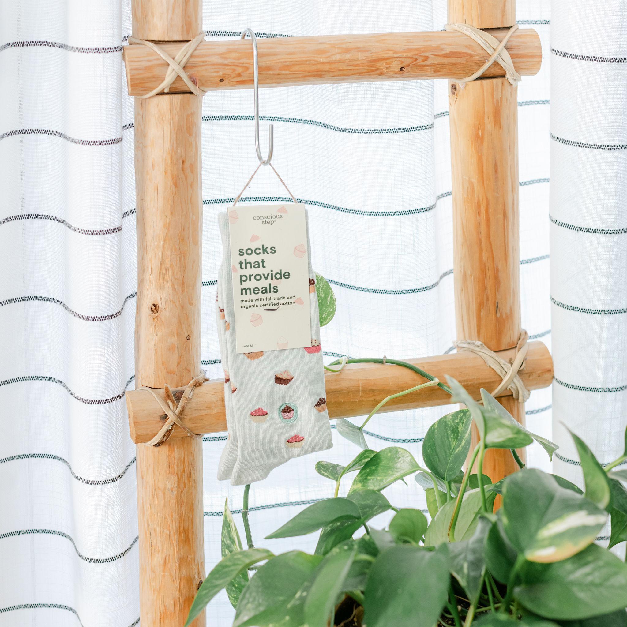 A package of socks with a cupcake pattern is hanging on a wooden display frame, with a plant in the background.