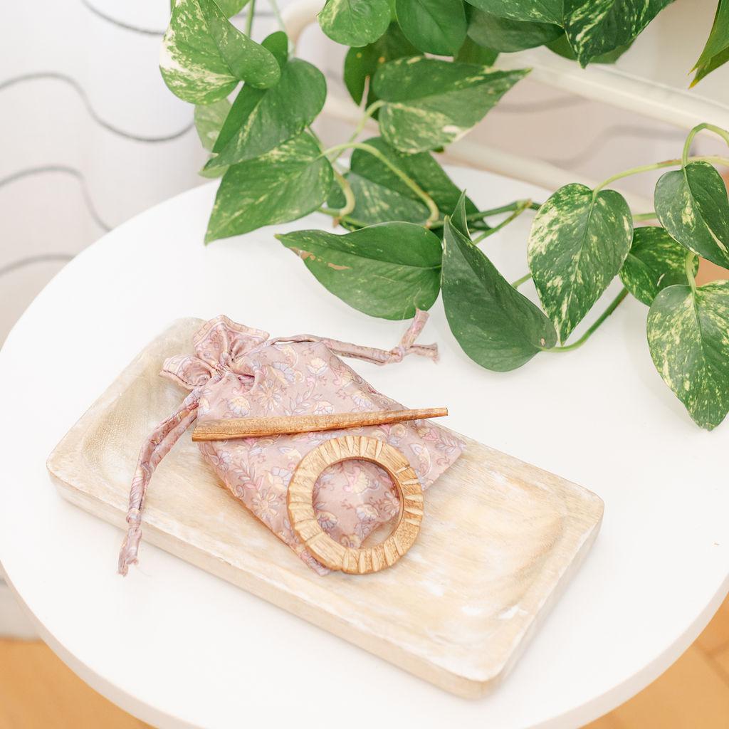 A mango wood hair slide with a ribbed design displayed on a white surface, accompanied by a matching wood hair stick, with a plant in the background.