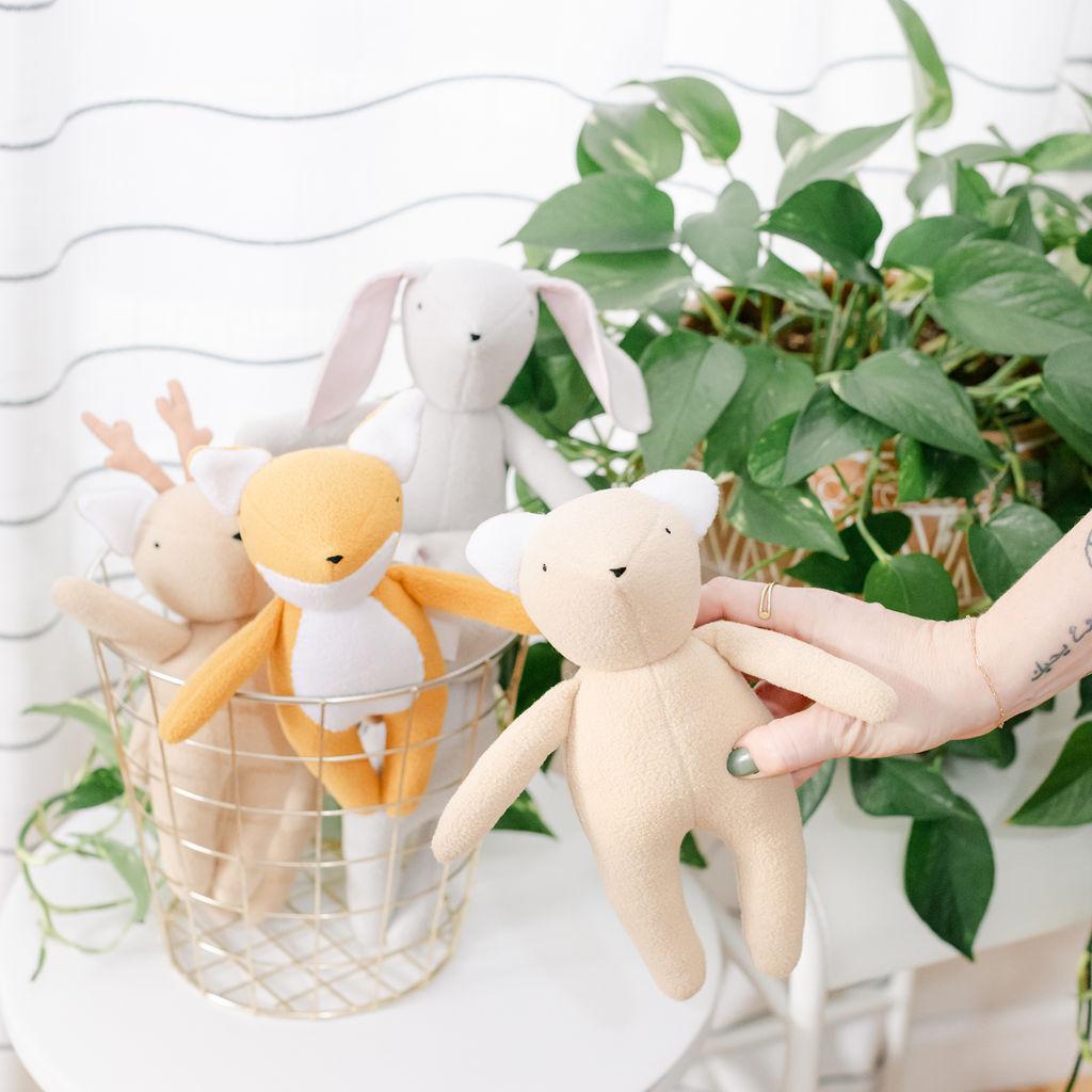 A beige teddy bear with a soft fleece body, displayed among other similar bears in a setting with green plants in the background.