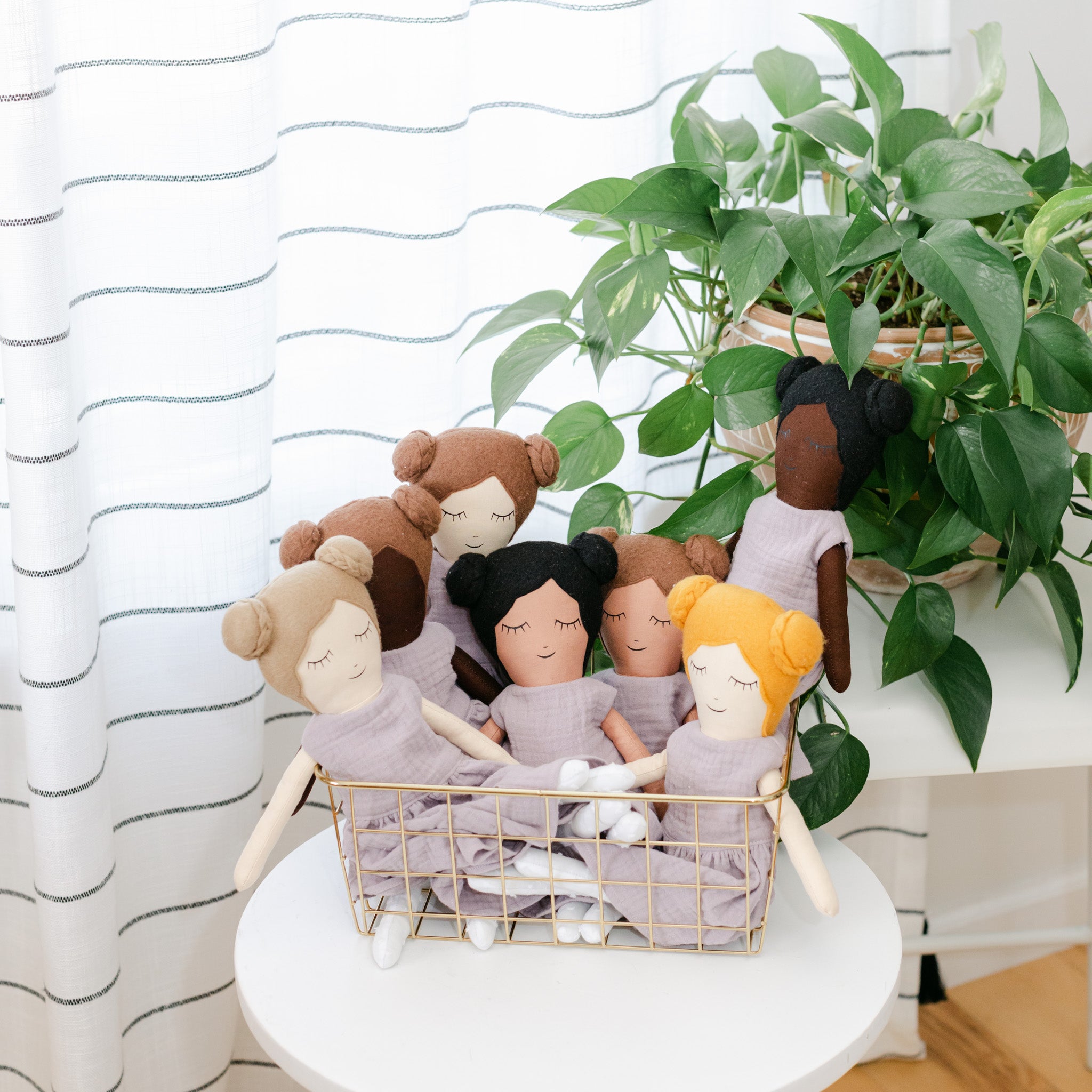 Stuffed multi-colored dolls in a basket on a white table with a plant in the backround with a striped curtain