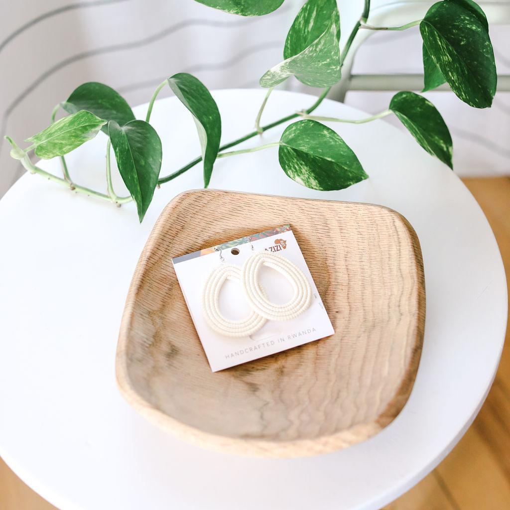 Wooden dish with a card featuring white woven hoop earrings on a white surface with green leaves.