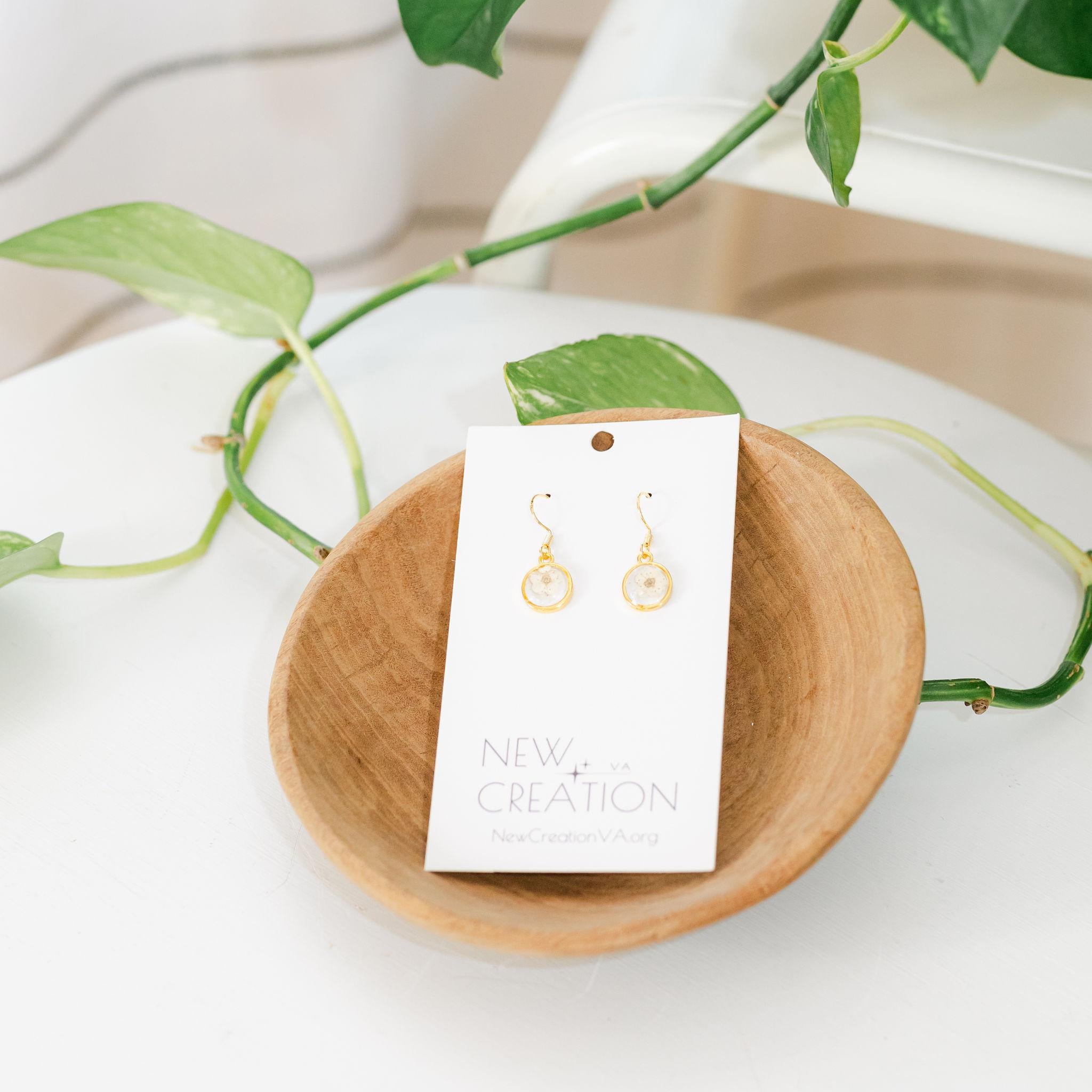 A pair of white wildflower earrings mounted on a presentation card, placed inside a wooden bowl with a plant in the background.