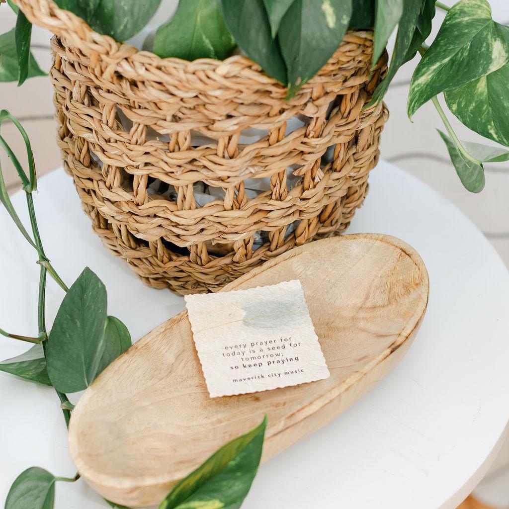 A small paper sign about prayer on a wooden plate by a wicker basket with a plant in the background
