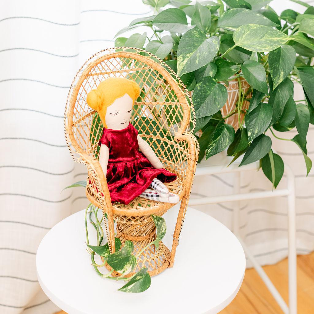 A doll with a maroon velvet dress presented sitting in a wicker chair against a backdrop of green plants.