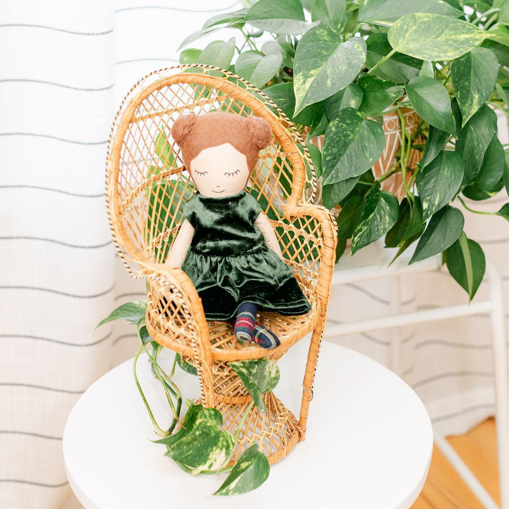 A doll with brown hair and buns wearing a green dress, sitting in a wicker chair with a plant in the background.