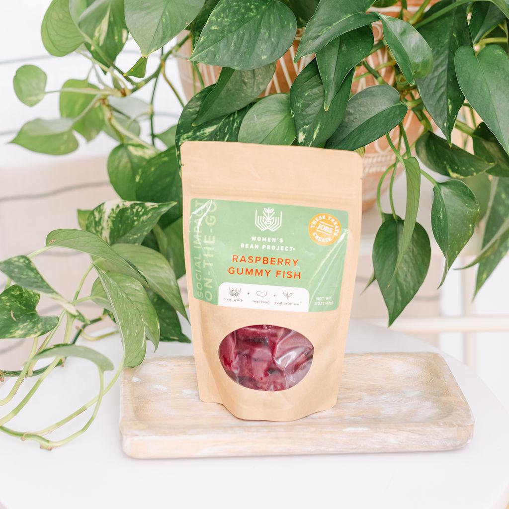 A resealable package of raspberry-flavored gummy fish snacks placed on a wooden surface with green leaves in the background.