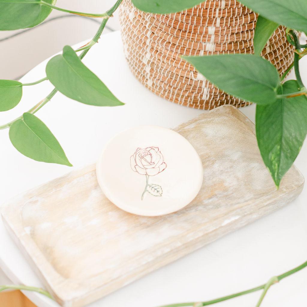 White ceramic dish with rose design on a wooden tray, surrounded by green leaves.