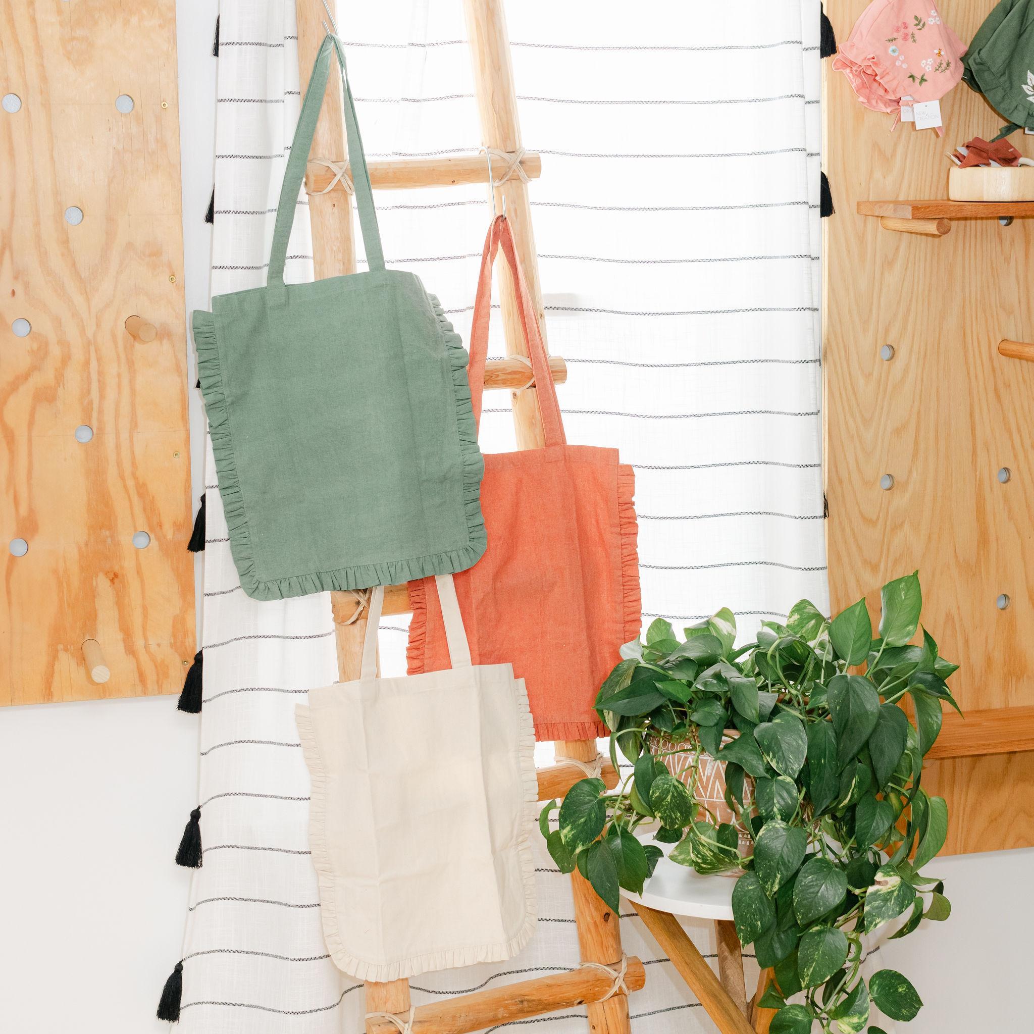 Three tote bags with ruffles hanging on a wooden ladder with a plant in the foreground.