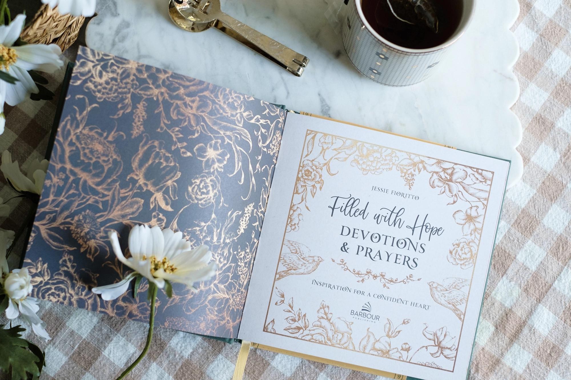 Open book with decorative cover and text on a checkered tablecloth with flowers and a cup.