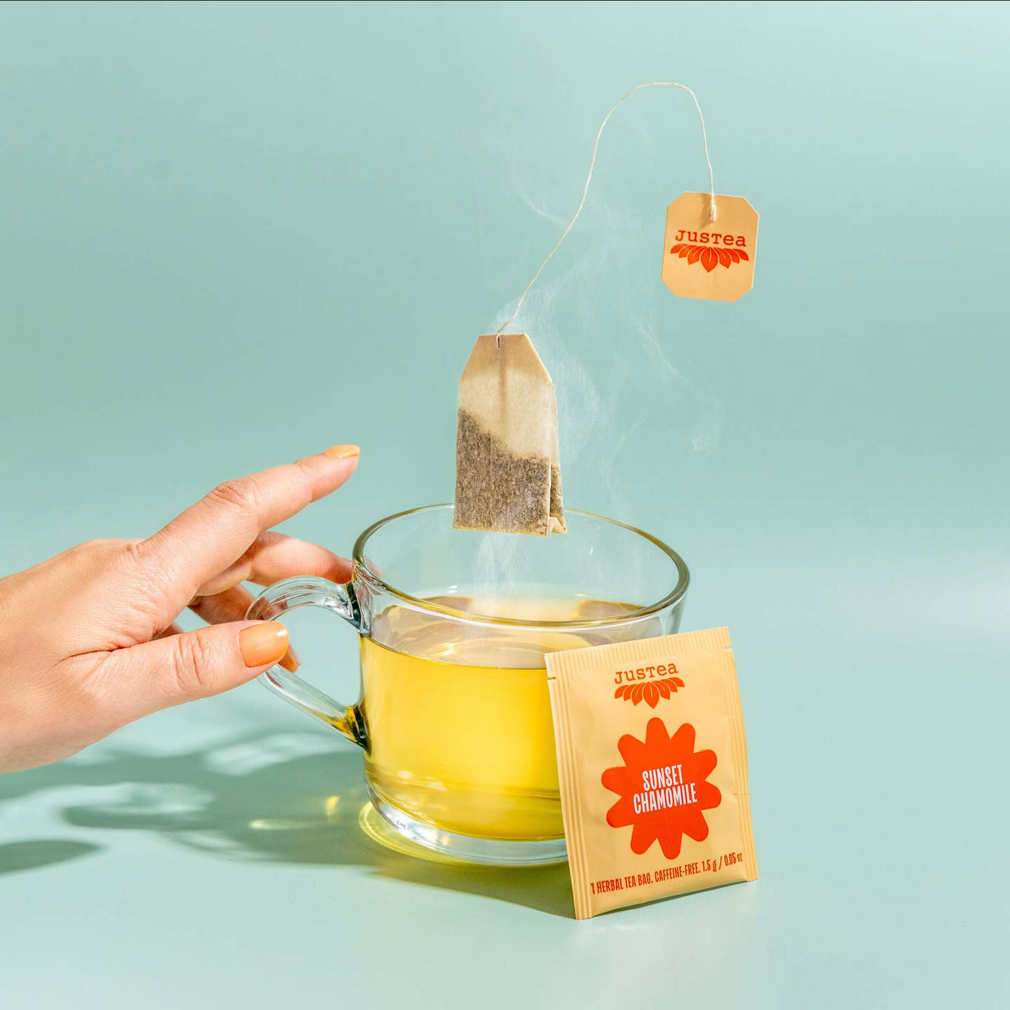 Hand holding a glass of yellow tea with a Just Tea sunset chamomile tea bag on a light blue background