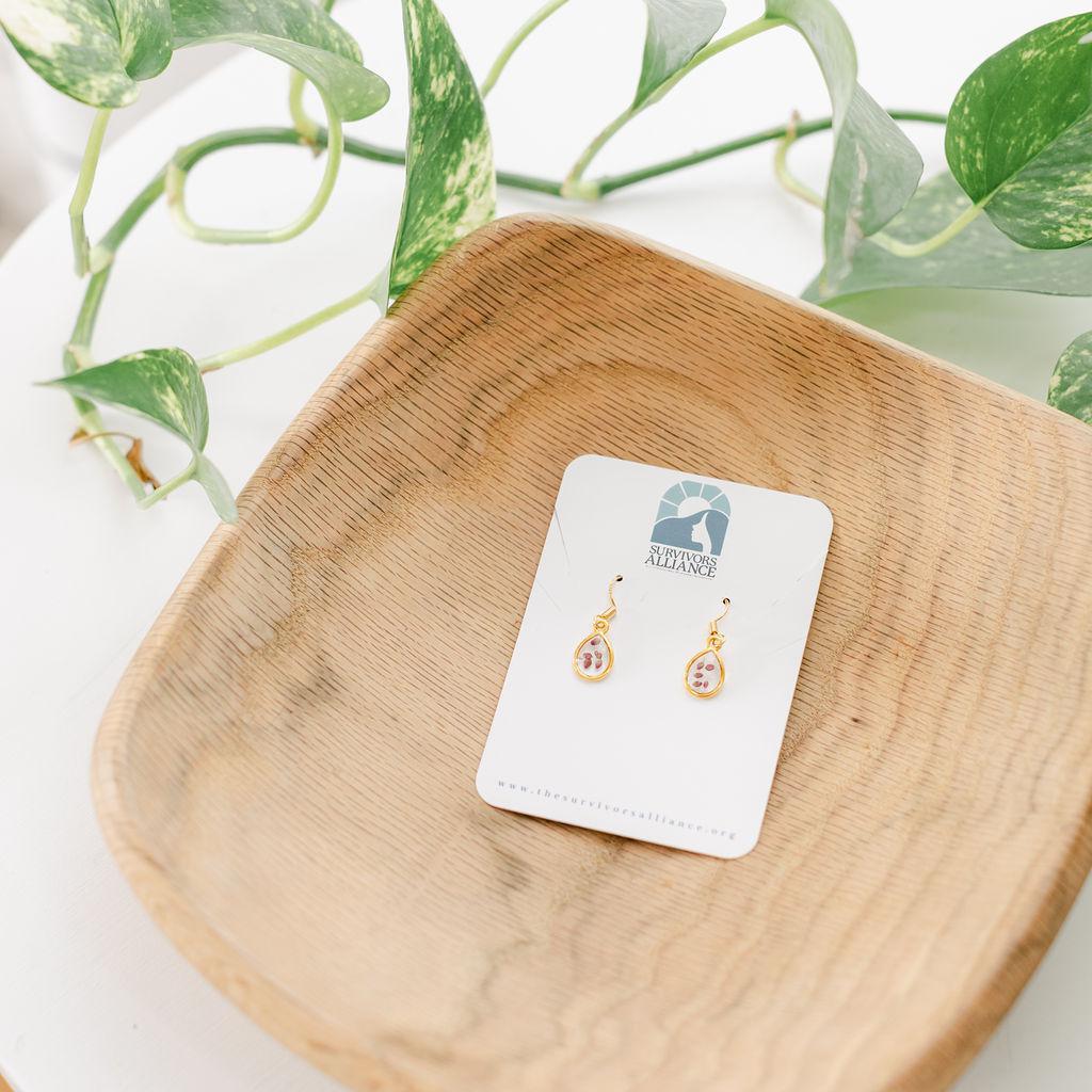 A pair of teardrop-shaped earrings with gold-plated, nickel-free, and hypoallergenic w/ pink/red flower buds in a clear resin, displayed on a wooden plate with a brand tag.