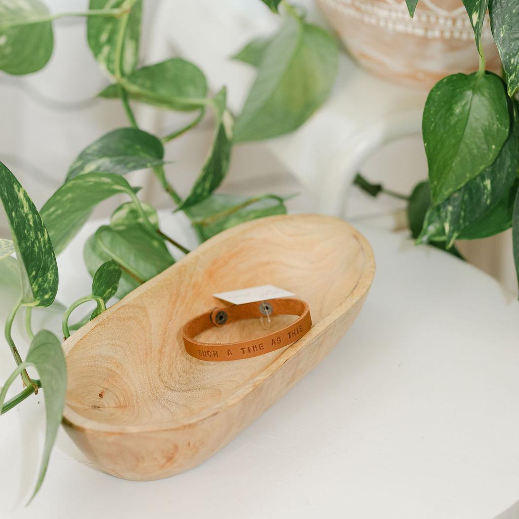 Wooden dish with a dark leather bracelet featuring the saying "for such a time as this", surrounded by green leaves on a light background