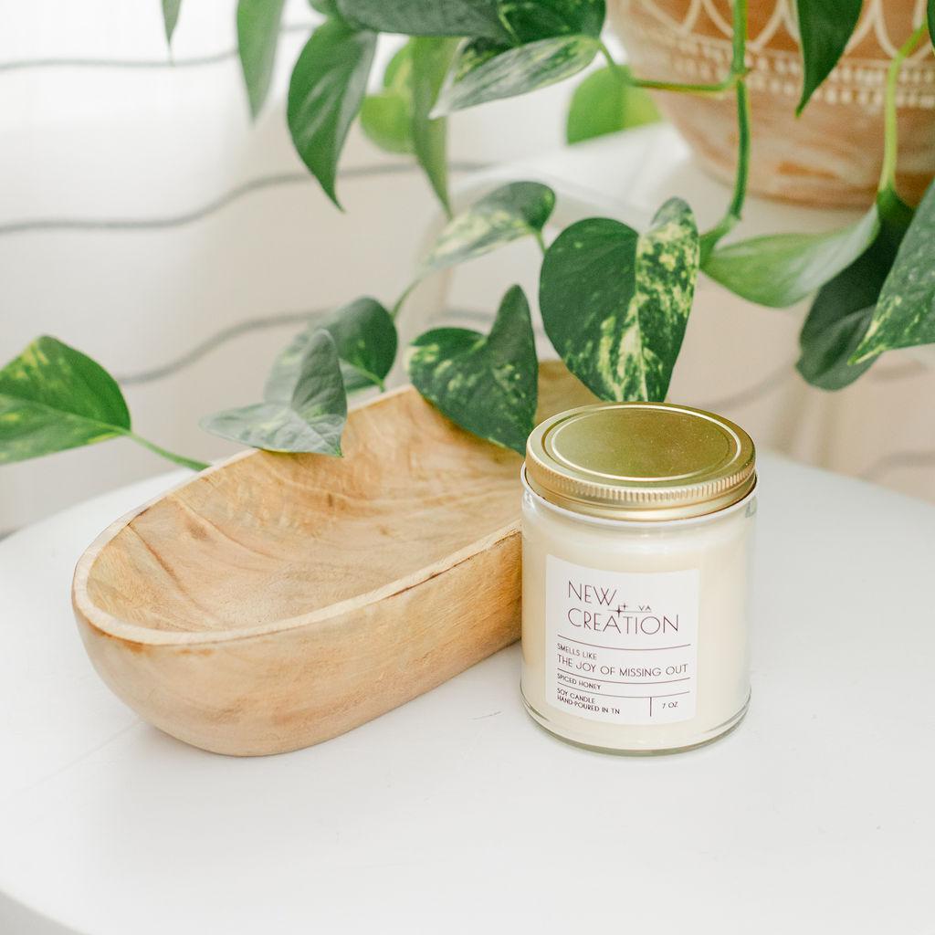 A hand-poured soy candle in a white container with a gold lid placed on a wooden surface with green leafy background.