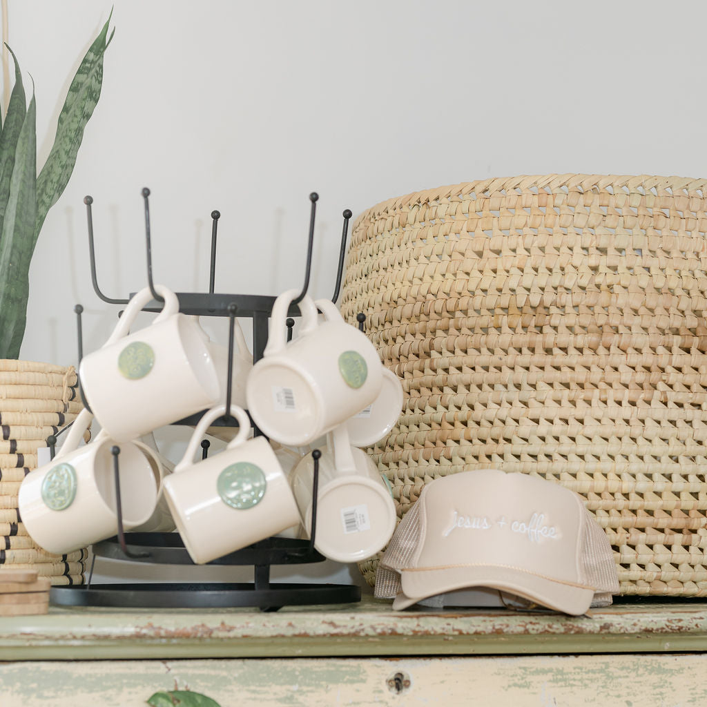 Set of white mugs with green logos on a stand next to a beige cap on a wooden surface.