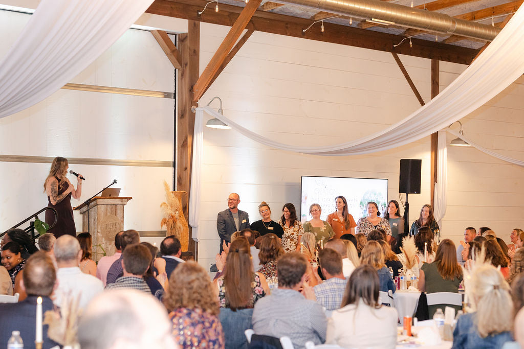 Woman speaking on a stage with an audience in a large room with wooden beams and white draped fabric.
