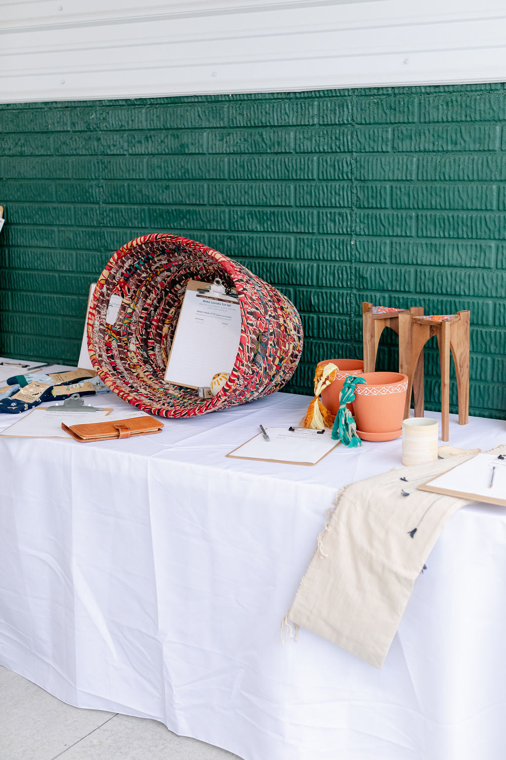 Table with various items including a colorful basket, terra cotta pots and a leather notepad against a green wall.