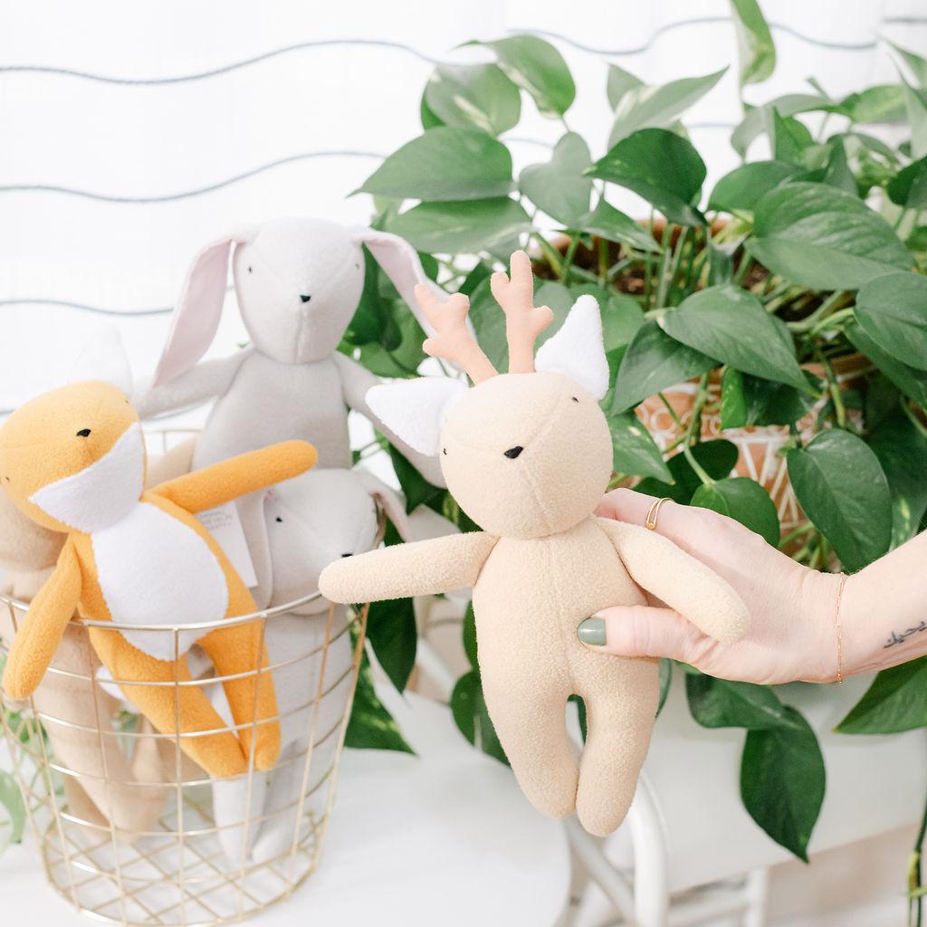 A beige plush toy deer with a white tail and antlers, being held by a person with green plants in the background.