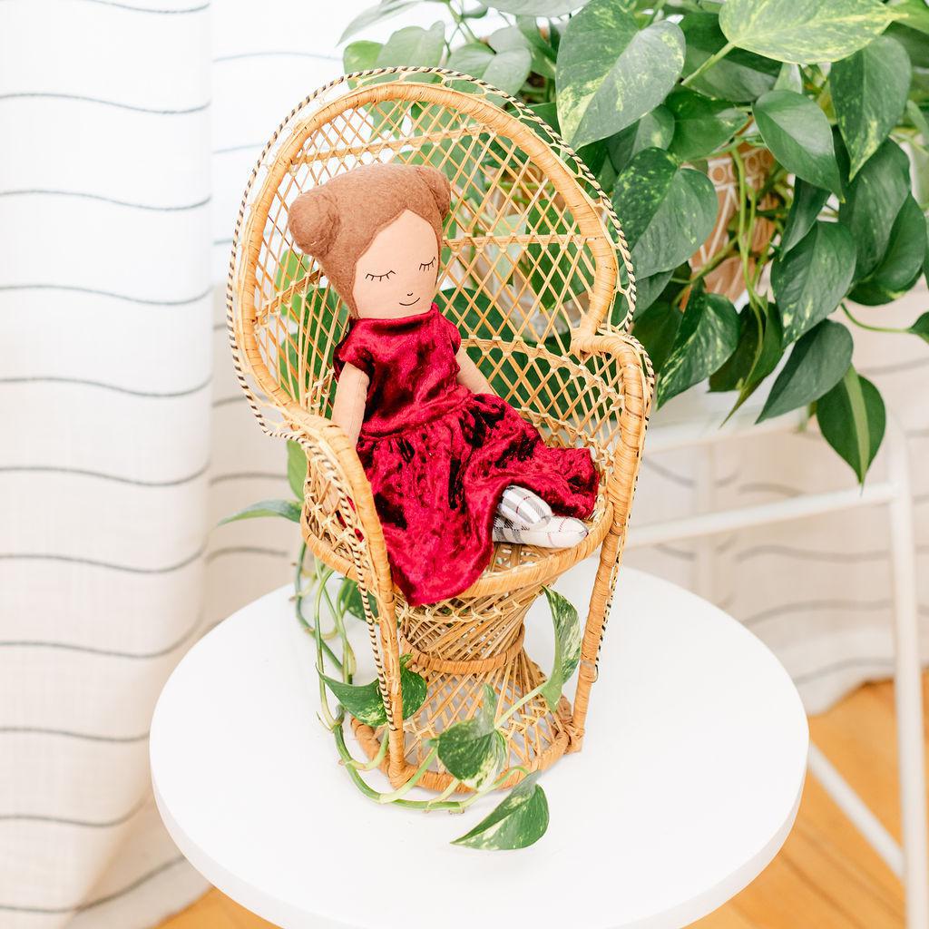 A doll with a maroon velvet dress presented sitting in a wicker chair against a backdrop of green plants.