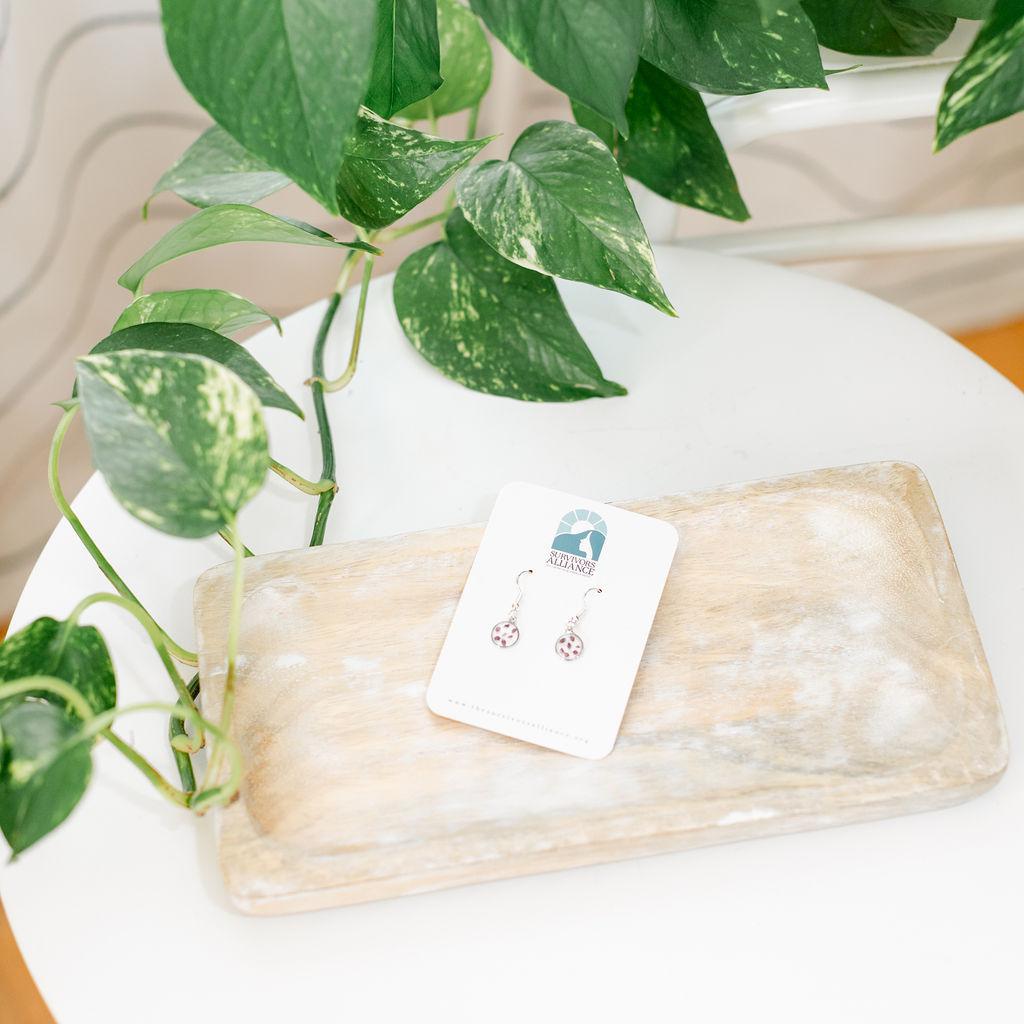 A pair of silver and pink flower bud resin earrings displayed on a flat surface with a plant in the background.