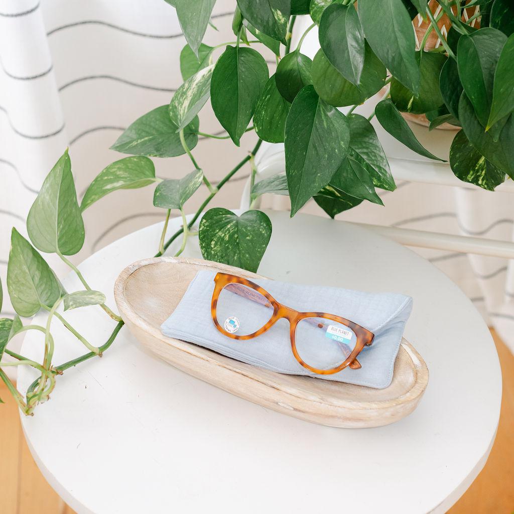 A pair of sunglasses placed inside a blue muslin sunglass case, which is resting on a wooden tray with a plant in the background.
