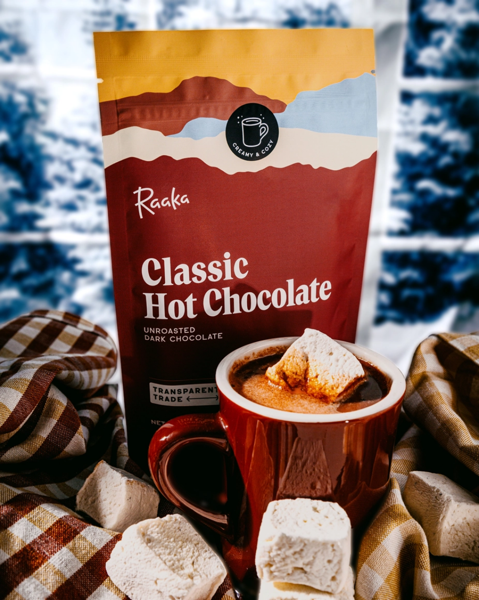 Bag of Raaka Classic Hot Chocolate with a mug of hot chocolate and marshmallows on a cozy blanket.