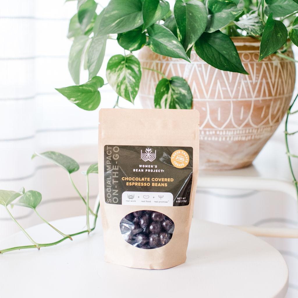 A resealable package of chocolate covered espresso beans placed on a white table with a plant in the background.