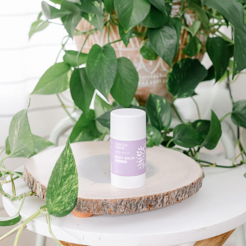 A container of Soothing Body Balm by Thistle Farms placed on a wooden surface with green plants in the background.