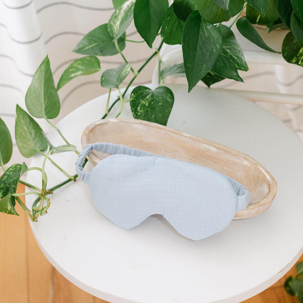 A blue muslin sleep mask placed on a white surface with green leaves in the background.