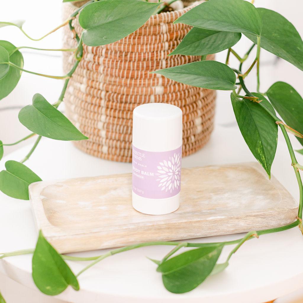 A container of Soothing Body Balm by Thistle Farms placed on a wooden surface with green plants in the background.