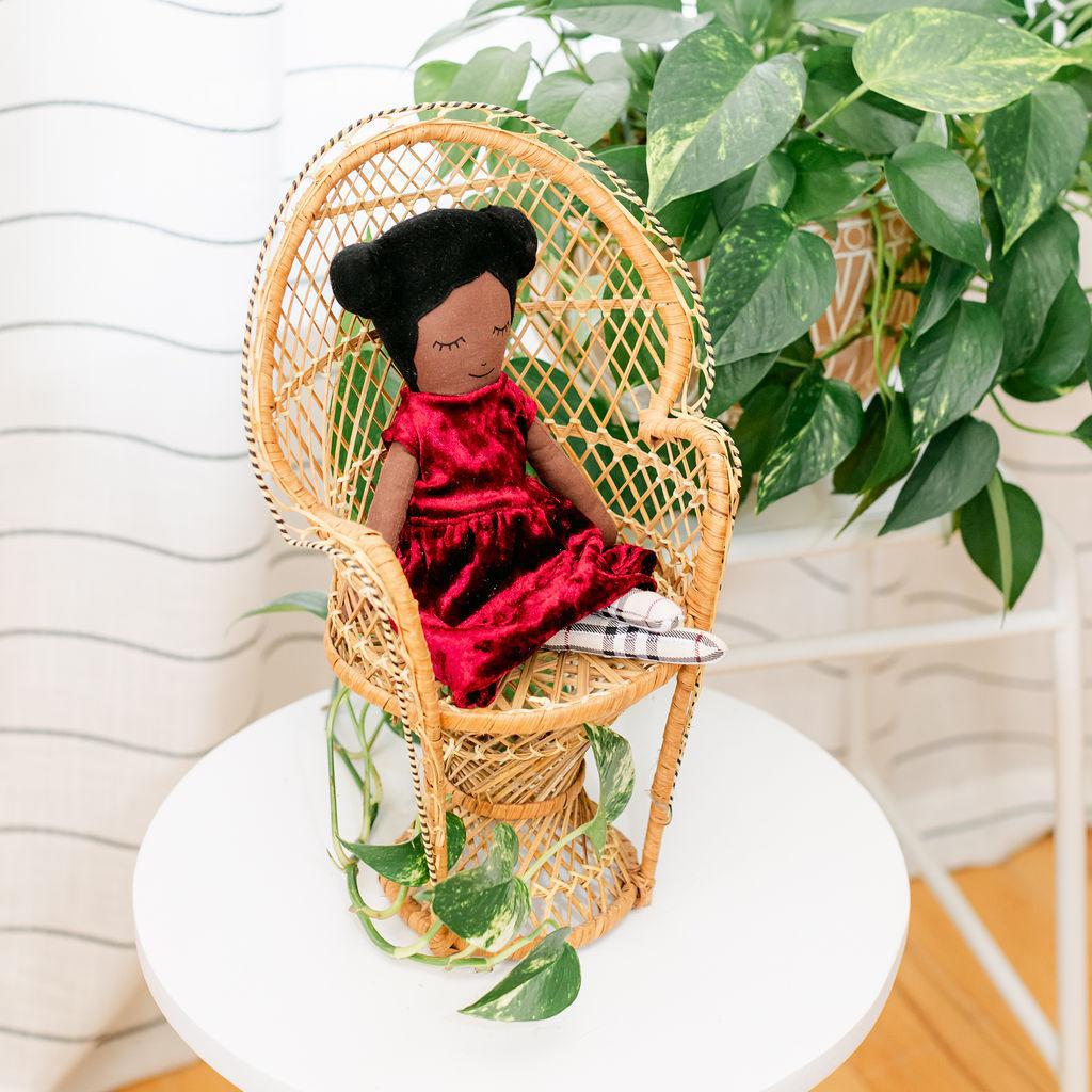 A doll with a maroon velvet dress presented sitting in a wicker chair against a backdrop of green plants.